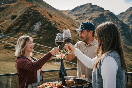 Weinprobe - Silvretta Montafon | © Silvretta Montafon - Vanessa Strauch Drei Personen stehen an einem Weinfass mit Käse und Brot und stoßen mit einem Glas Rotwein an. | © Silvretta Montafon - Vanessa Strauch