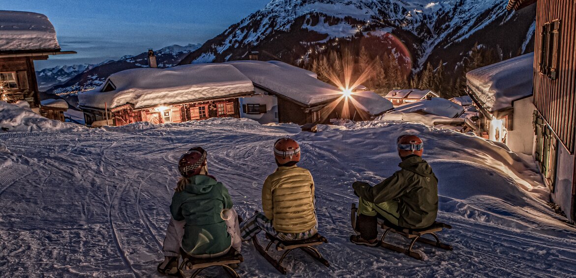 Drei Personen sitzen auf einem Schlitten in einem Bergdorf am Abend in der Silvretta Montafon. | © Silvretta Montafon - Daniel Zangerl