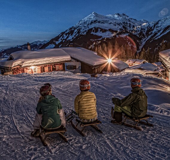 Nachtrodeln - Silvretta Montafon | © Silvretta Montafon - Daniel Zangerl Drei Personen sitzen auf einem Schlitten in einem Bergdorf am Abend in der Silvretta Montafon. | © Silvretta Montafon - Daniel Zangerl