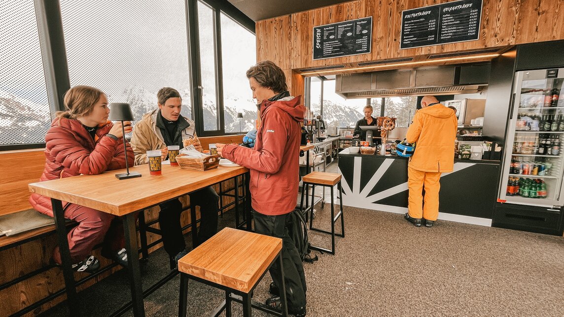 Innenraum Sonnen Barga - Silvretta Montafon | © Silvretta Montafon - Vanessa Strauch Drei Personen in der Sonnenbarga an einem Stehtisch und eine Person bestellt etwas an der Theke. | © Silvretta Montafon - Vanessa Strauch