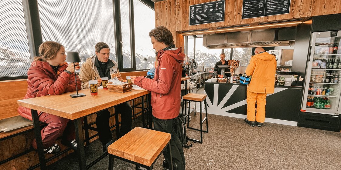 Innenraum Sonnen Barga - Silvretta Montafon | © Silvretta Montafon - Vanessa Strauch Drei Personen in der Sonnenbarga an einem Stehtisch und eine Person bestellt etwas an der Theke. | © Silvretta Montafon - Vanessa Strauch