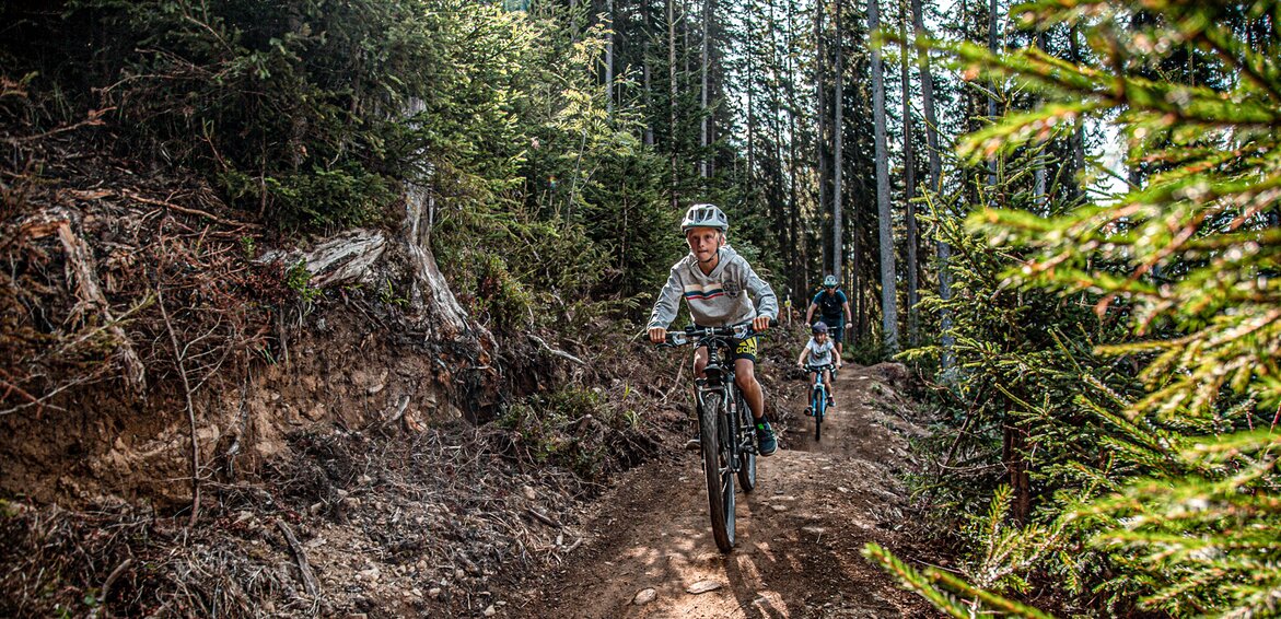 Eine Junge im Vordergrund und zwei Personen im Hintergund fahren durch den Wald im Bikepark am Hochjoch in der Silvretta Montafon.  | © Silvretta Montafon - Friederike Weber