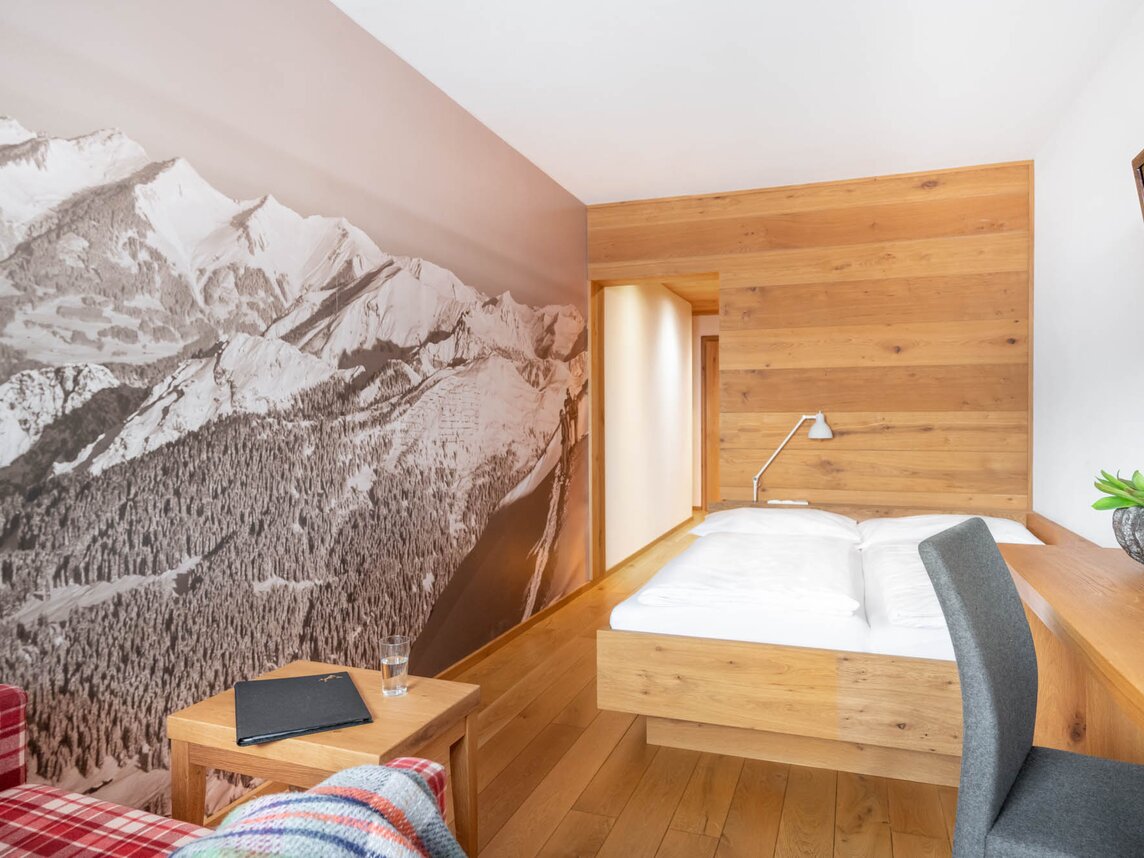 Doppelzimmer im Sporthotel Silvretta Montafon | © Silvretta Montafon - blateral Ein Doppelbett mit Holzrahmen vor einer Holzwand und eine rot-karierter Couch | © Silvretta Montafon - blateral